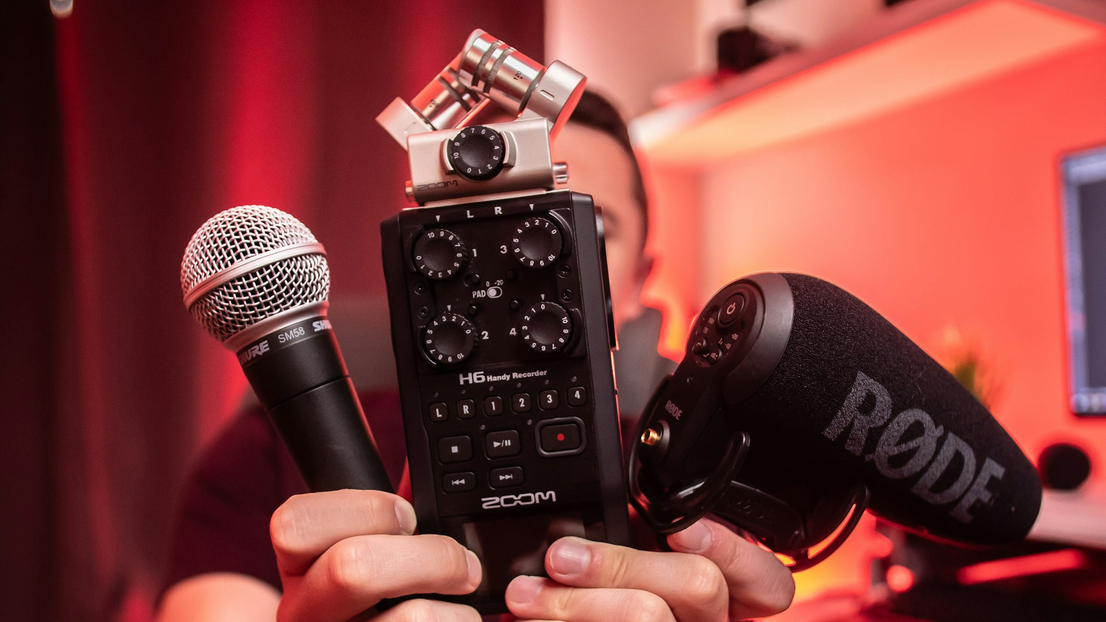 Hands holding microphones and a portable audio recorder against a red background.